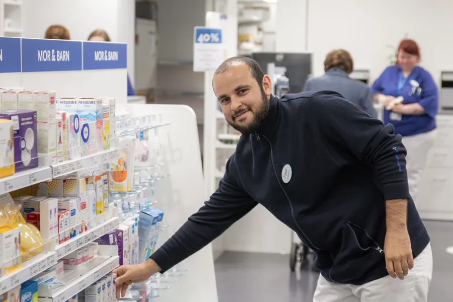 Faisal rydder i hyllene på apoteket mens han smiler til kamera