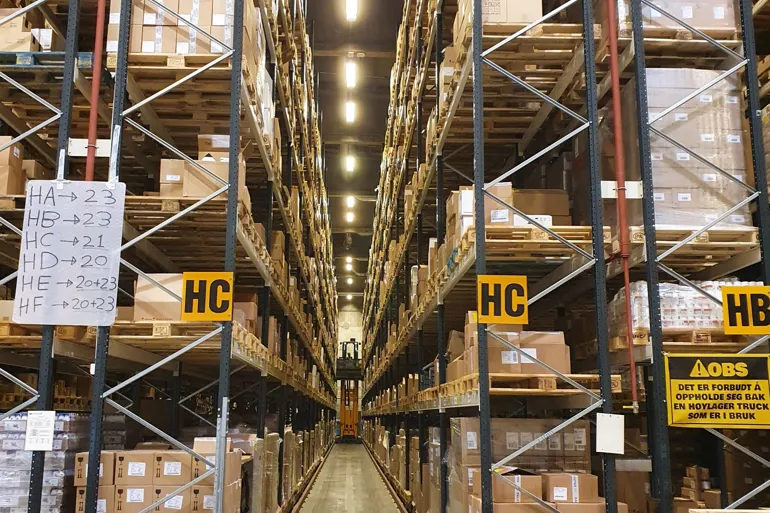 Bilde av lagerhyller i et stort lager. En truck kommer kjørende i enden av lagerhyllene.