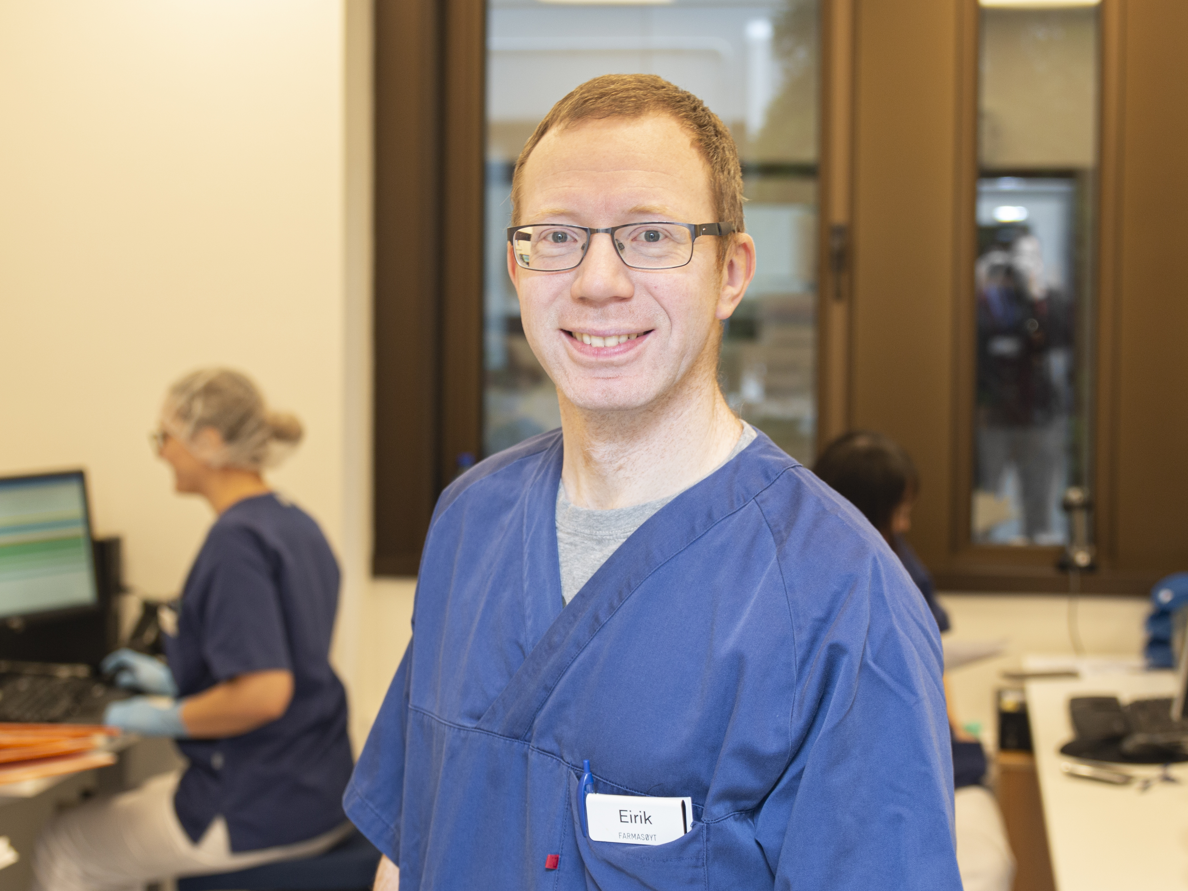 Eirk Svandal smiler til kamera, mens en kollega jobber ved PCen i bakgrunnen