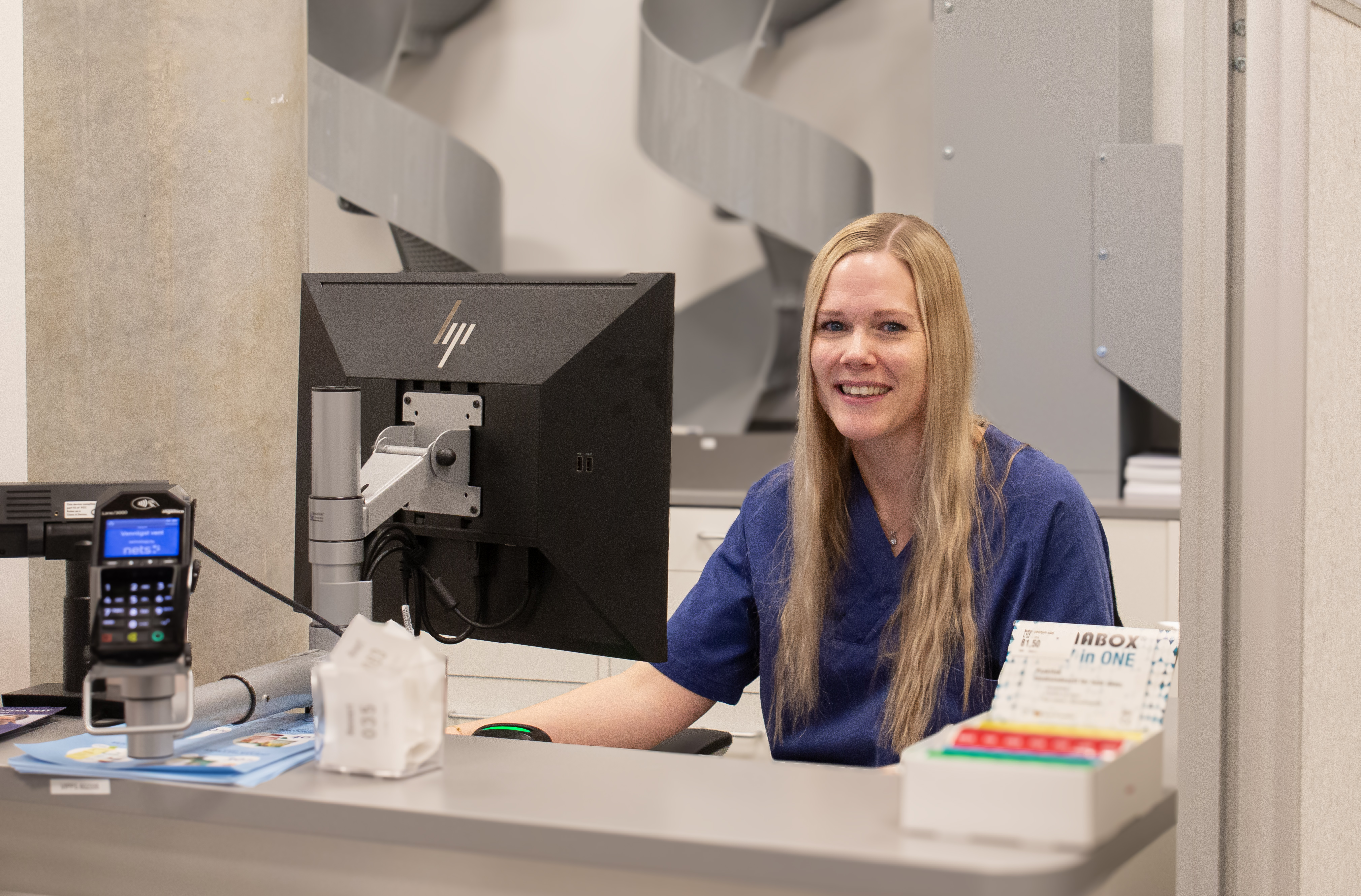 En kvinne sitter i resepturen klar for å ta imot pasienter som skal hente resepten sin. Hun sitter bak en pult med en PC-skjerm.
