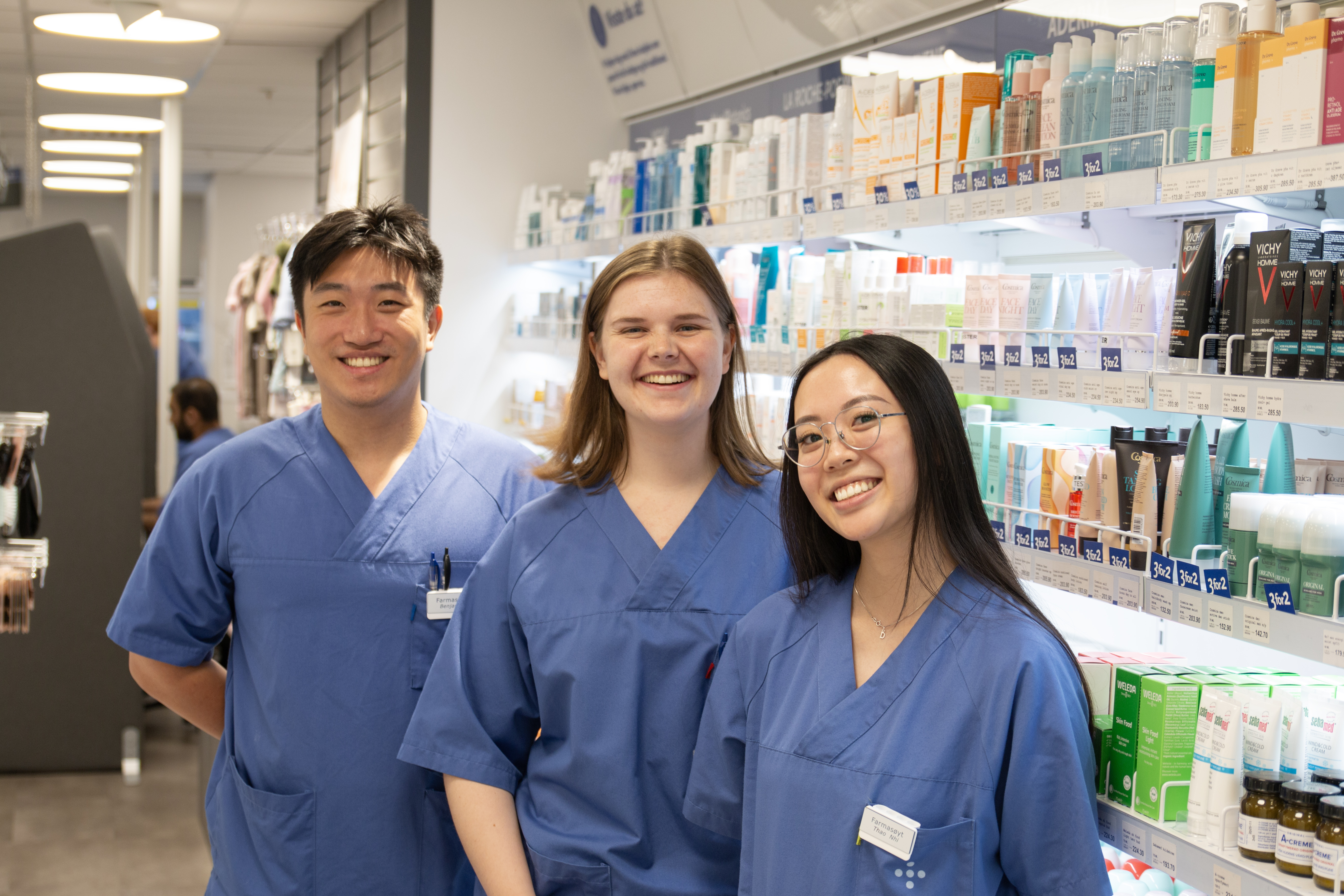 Tre unge farmasøyter står ved en hylle med varer og smiler til kamera.